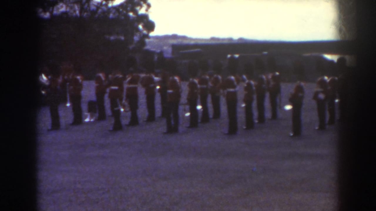 1938 Quebec home-movie frame showing a marching band in red uniforms lined up outdoors on a gravel field.