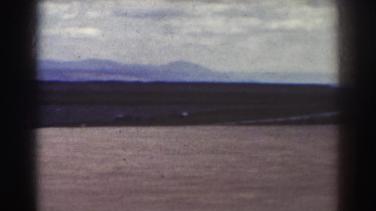 1938 Quebec home-movie frame showing an empty dirt field, low horizon, distant hills, and overcast sky through dark film borders.