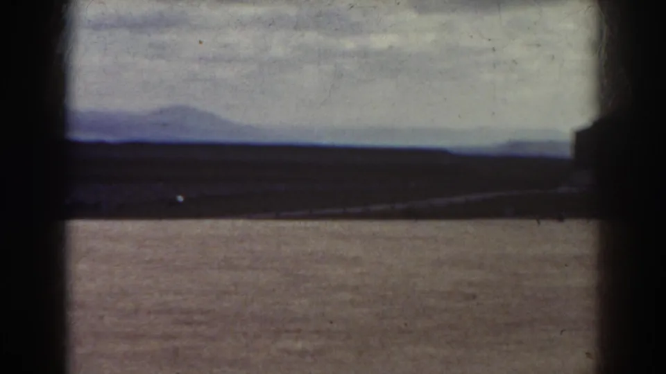 1938 Quebec landscape frame with open dirt ground, distant hills, overcast sky, and a dark building edge entering at right.