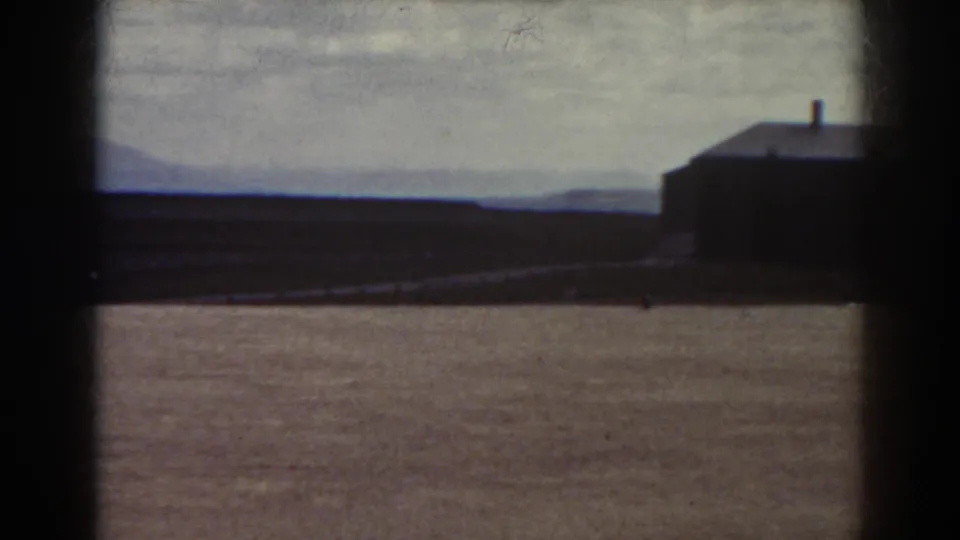 1938 Quebec home-movie frame showing a low dark building near an empty field, distant hills, and a cloudy sky.
