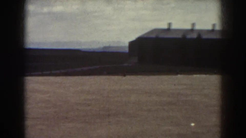 1938 Quebec frame showing a dark low building with roof vents, open dirt field, distant hills, and overcast sky.