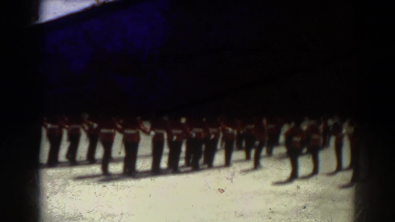 1938 Quebec home-movie frame showing soldiers in red tunics and tall dark hats standing at attention on a parade ground.