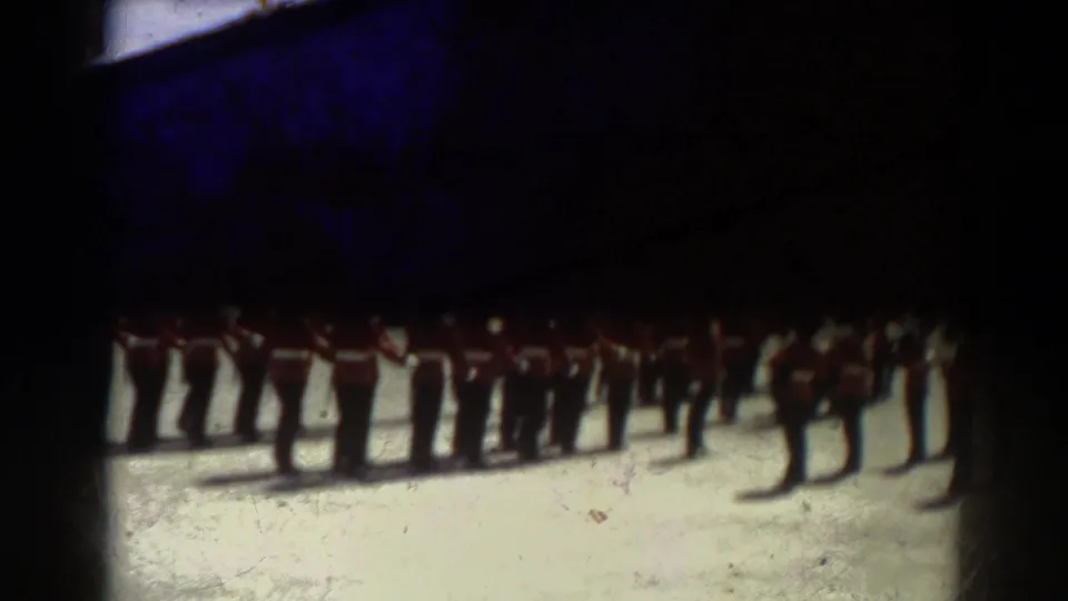 1938 Quebec home-movie frame showing soldiers in red tunics and tall dark hats standing at attention on a parade ground.