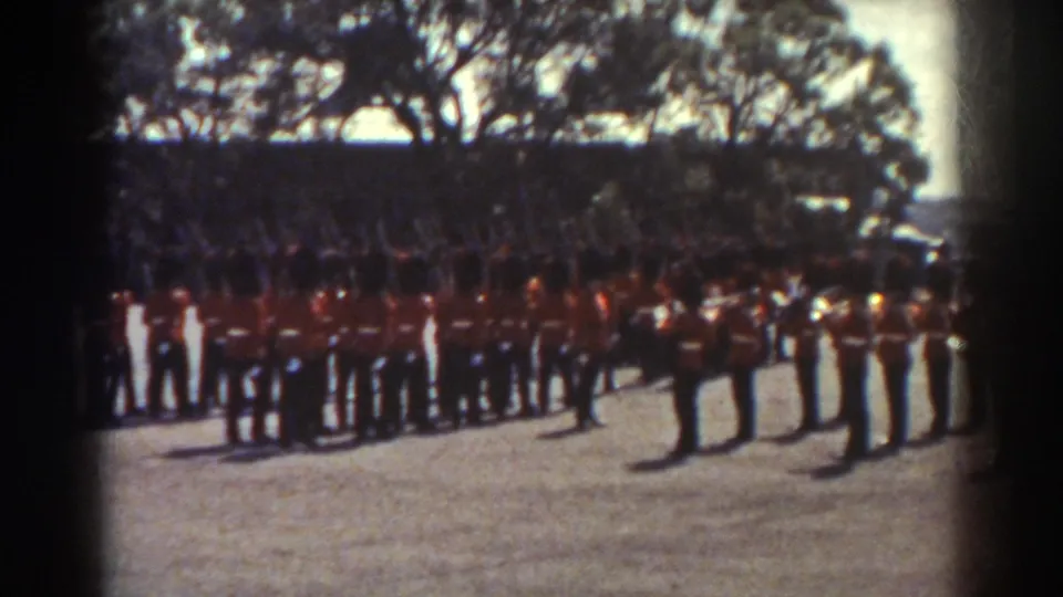 Red-uniformed soldiers in Quebec stand in ceremonial formation, with trees and shaded background figures behind them.