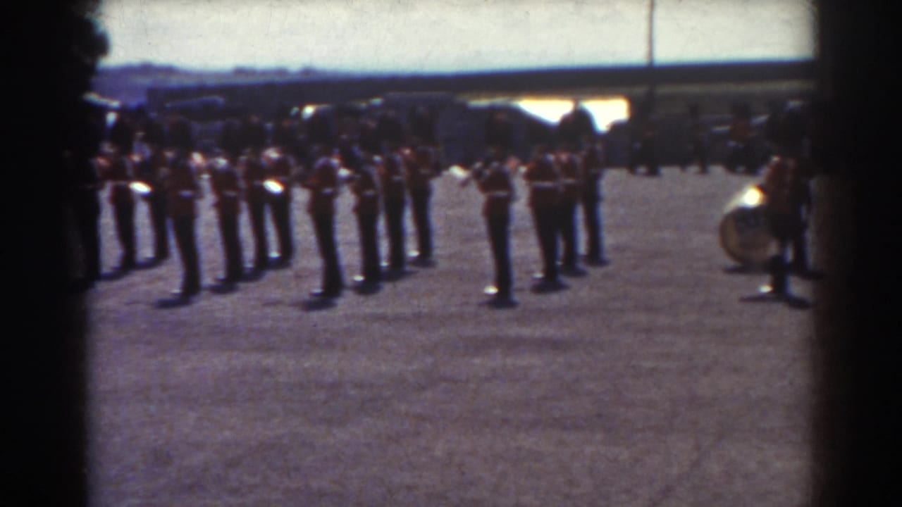 Uniformed soldiers stand in formation on a gravel field with a drum partly visible at right.