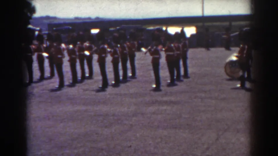 Uniformed soldiers in Quebec stand in formation on a gravel field, with dark framing at the sides and a drum at right.