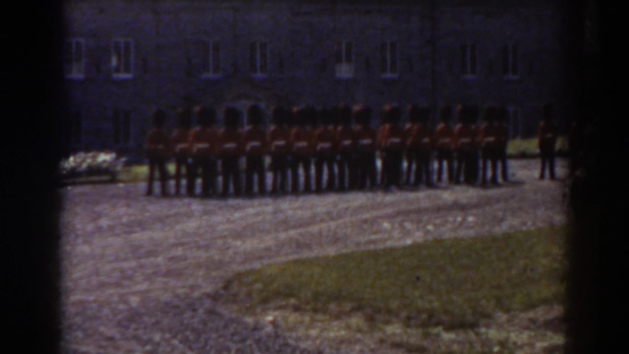 1938 Quebec home-movie frame showing red-coated soldiers with tall dark hats lined up before a stone building.