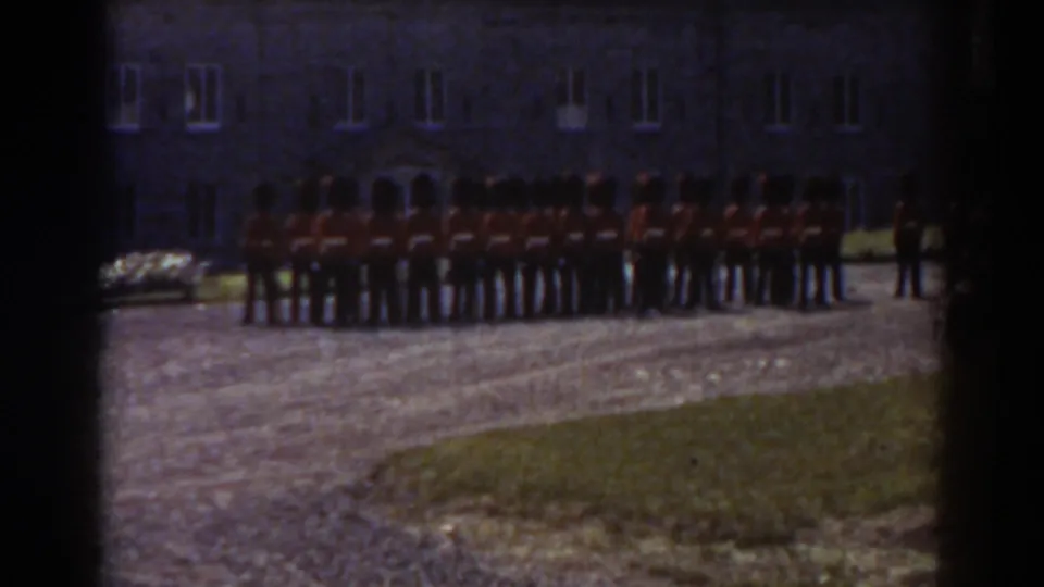 1938 Quebec home-movie frame showing red-coated soldiers with tall dark hats lined up before a stone building.