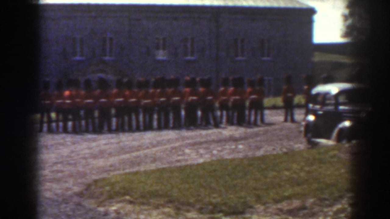 1938 Quebec frame with soldiers in red coats lined up near a stone building and a period car entering at right.
