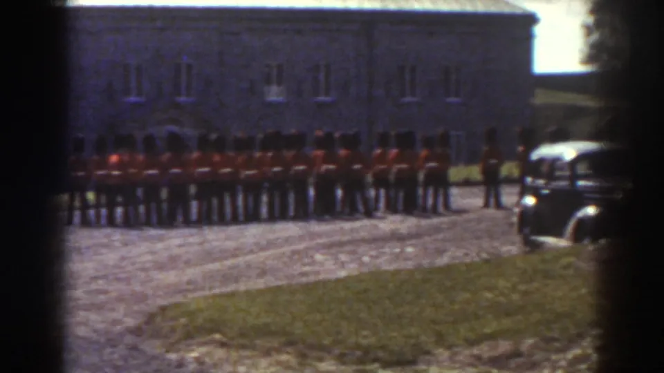 1938 Quebec frame with soldiers in red coats lined up near a stone building and a period car entering at right.