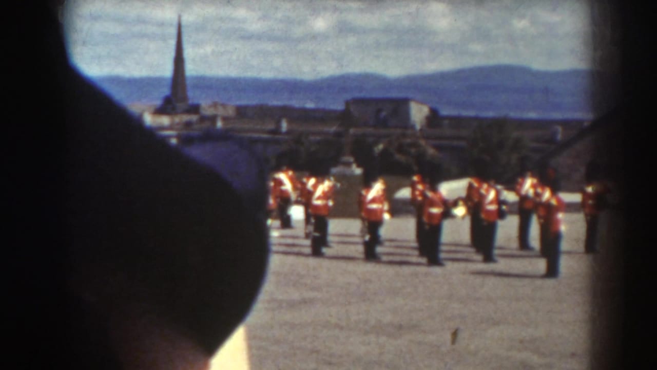 A line of red-uniformed soldiers stands near stone buildings, partly blocked by a dark hat.