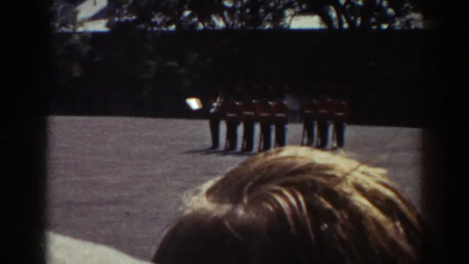 1938 Quebec frame showing soldiers in red uniforms at attention across a sunlit courtyard, with a spectator's head in front.