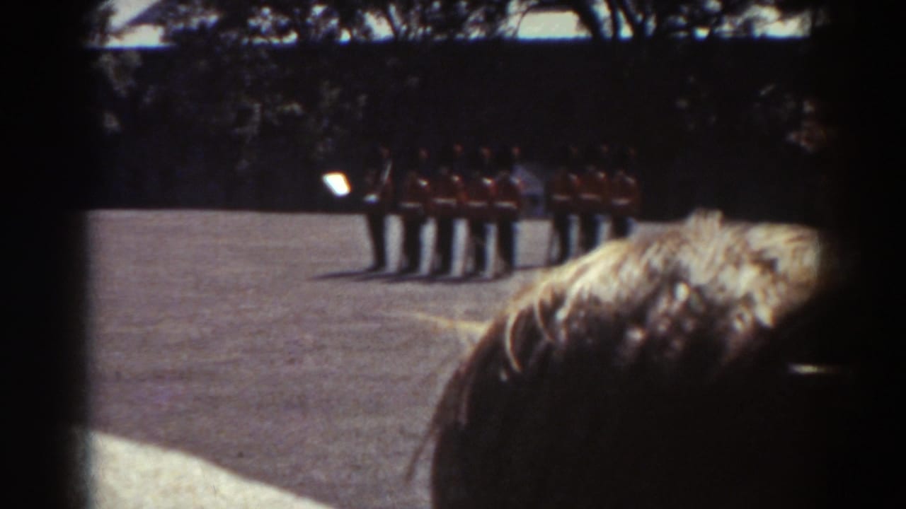 In 1938 Quebec footage, a foreground spectator watches red-coated guards in tall dark hats standing in formation on a sunny paved courtyard.