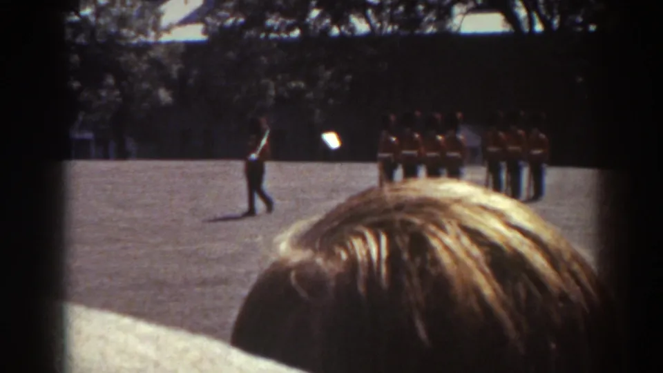 In the Quebec home-movie scene, one uniformed guard marches across open pavement while other red-coated guards remain lined up behind him.
