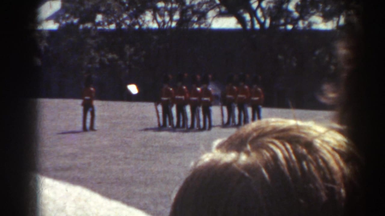 1938 Quebec home-movie frame showing spectators watching red-uniformed men in formation across a sunny open ground.