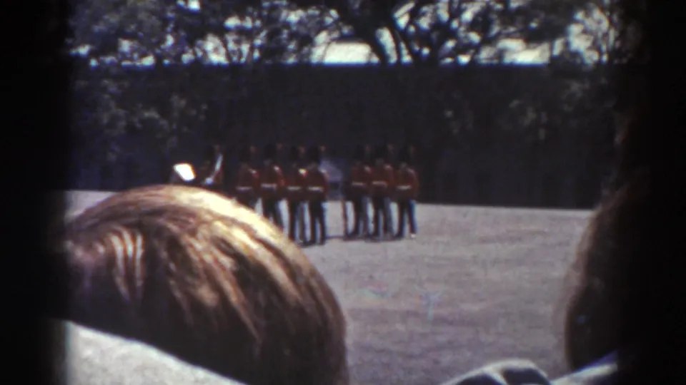 Handheld 8mm view of a Quebec military-style ceremony, with a spectator's head low in frame and uniformed men across the ground.