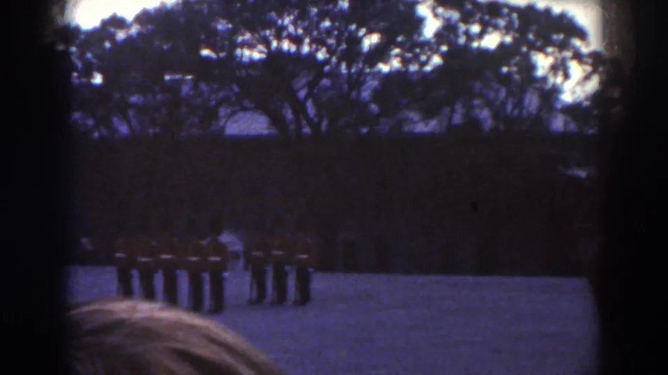 Red-coated soldiers stand or move in a formal outdoor drill in Quebec, filmed from behind onlookers on 8mm color stock.
