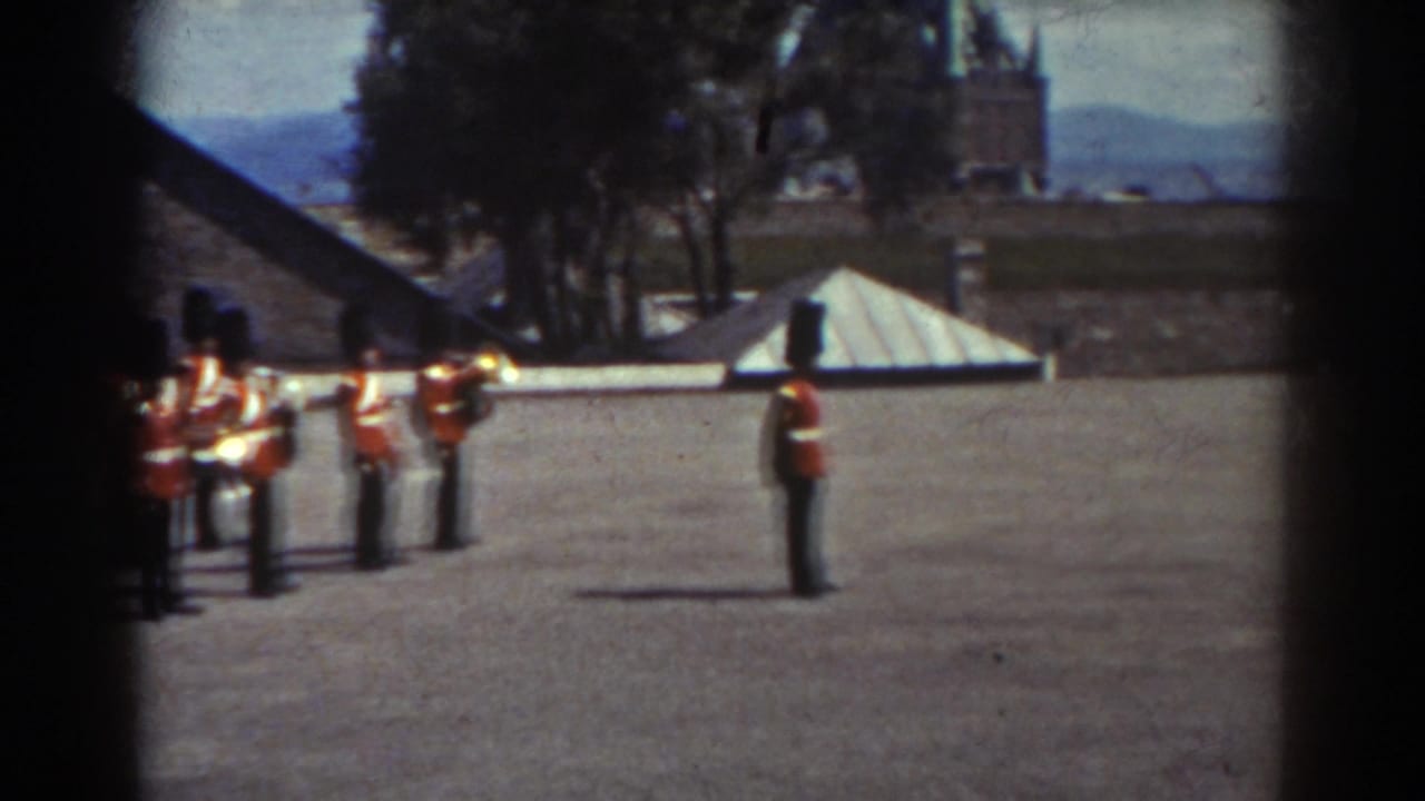 Several guards with rifles stand near stone buildings and trees.