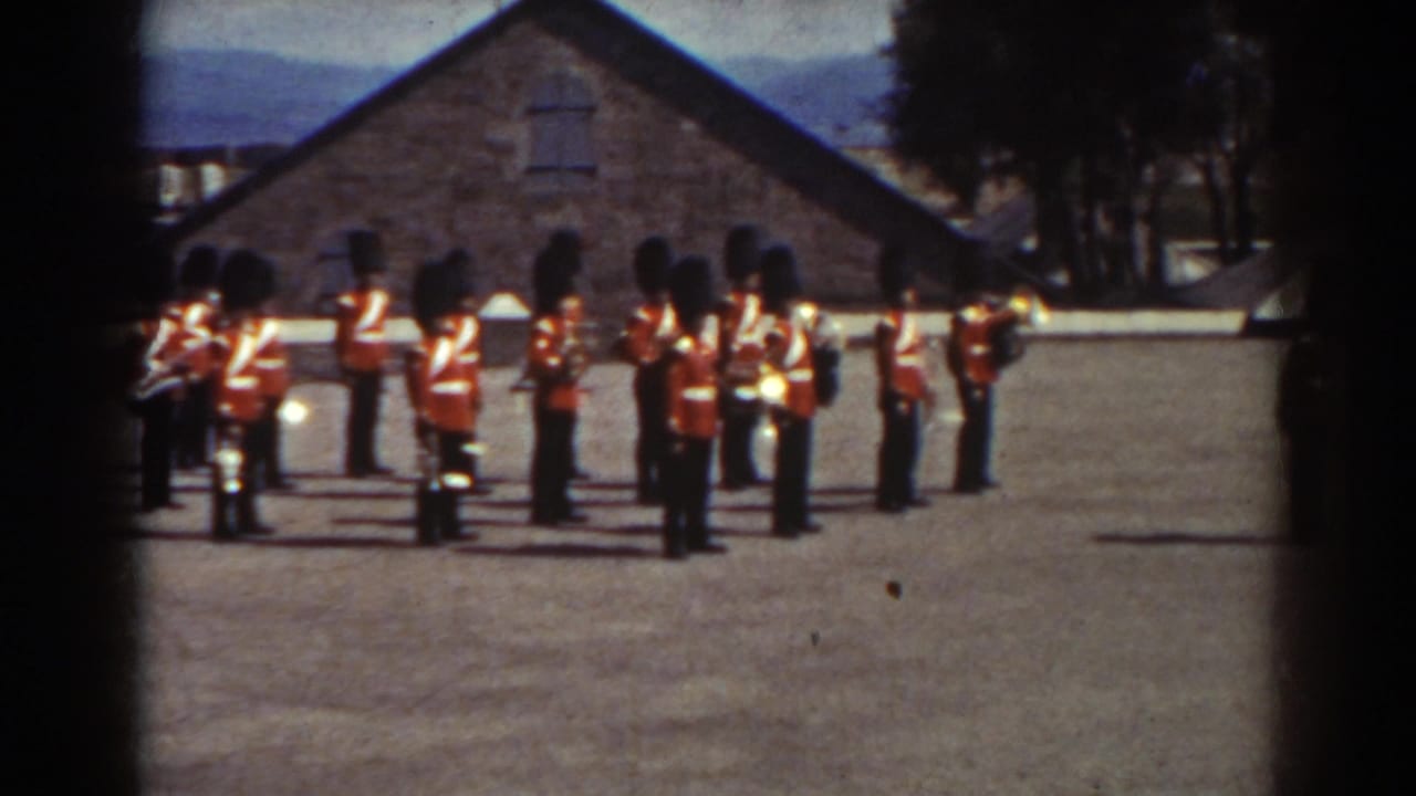 In 1938 Quebec footage, uniformed musicians in red jackets and tall black hats stand with brass instruments before stone buildings and distant hills.