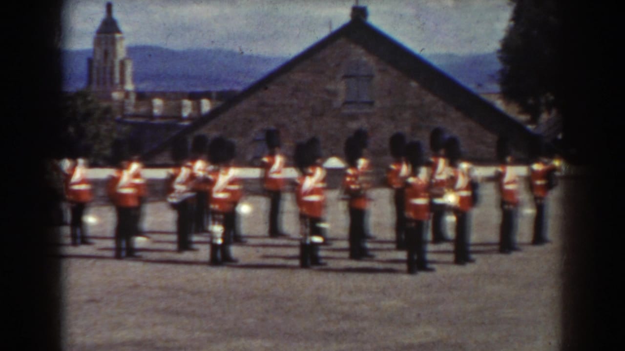 Marching band in red uniforms stands across a courtyard with a steeple behind.