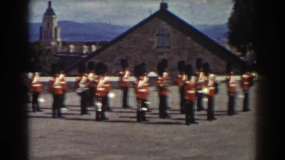 The band remains lined up in the Quebec courtyard, with brass instruments, black hats, stone architecture, and a church steeple visible behind them.