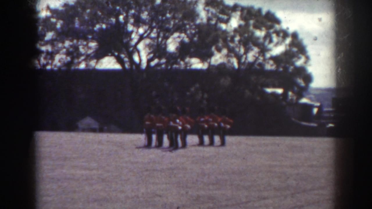 1938 Quebec 8mm frame showing a distant red-uniformed band lined up on grass before trees and a dark wall.