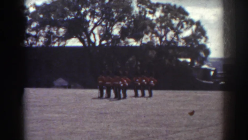 Distant 1938 Quebec home-movie view of red-uniformed band members standing in formation on a grass field.