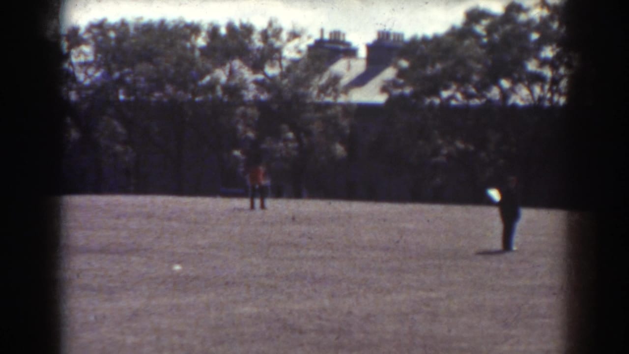 1938 Quebec 8mm frame showing two distant figures crossing an open field with trees and houses in the background.