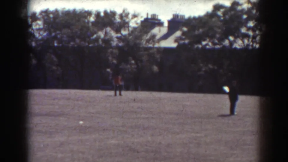 1938 Quebec 8mm frame showing two distant figures crossing an open field with trees and houses in the background.