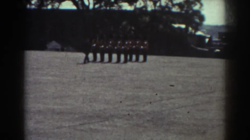 Distant Quebec 1938 frame of uniformed men standing in a line on a grassy field, with trees and dark equipment or vehicles behind them.