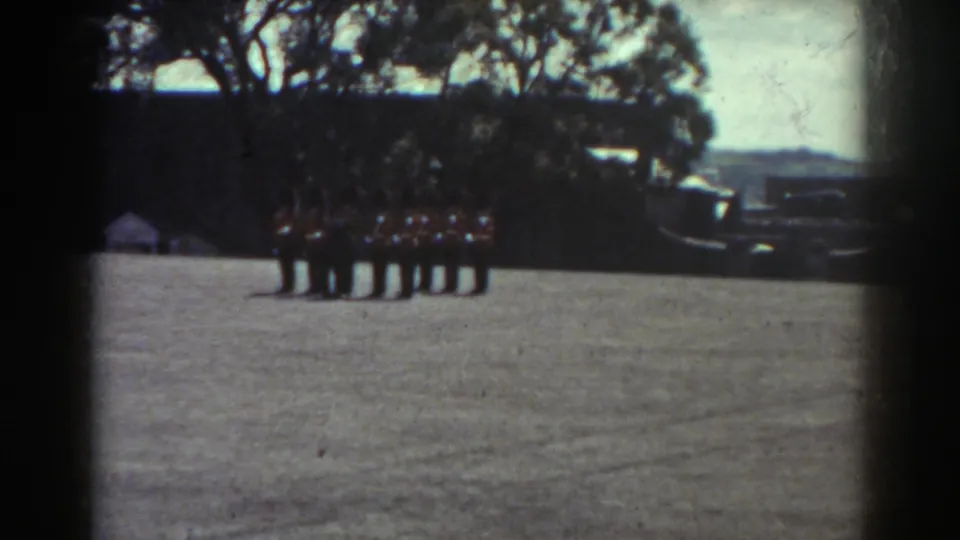Final Quebec 1938 frame of dark-uniformed men standing in formation on a field, with trees and indistinct vehicles or equipment behind them.