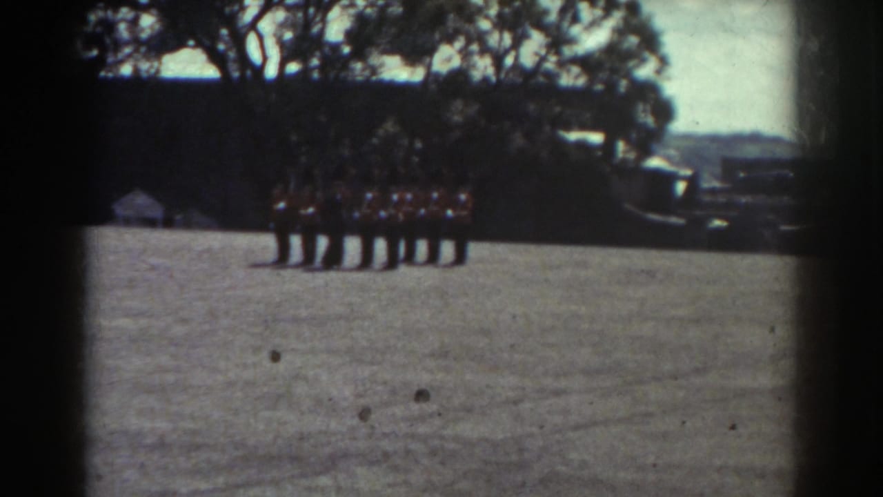 1938 Quebec home-movie frame showing red-jacketed ceremonial guards in formation on a gravel courtyard near trees and masonry buildings.