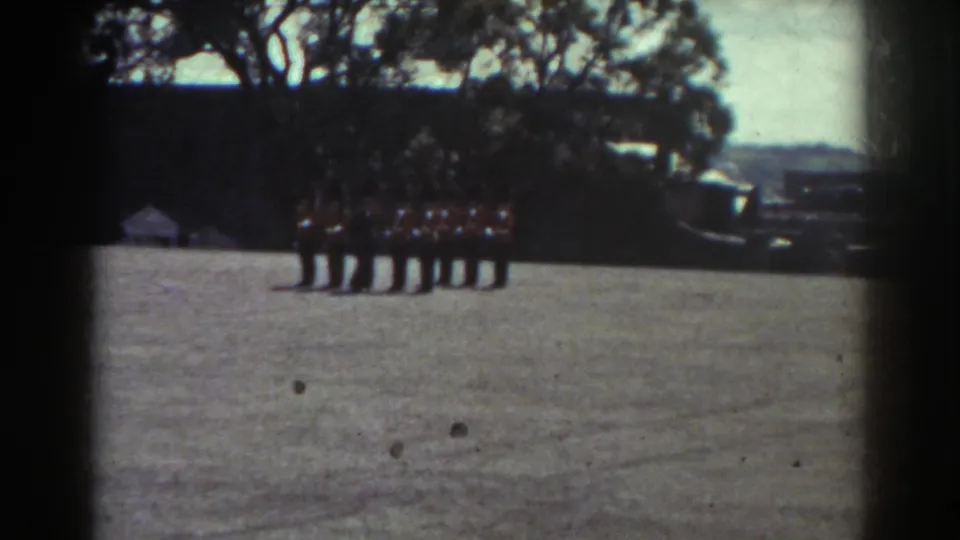 1938 Quebec home-movie frame showing red-jacketed ceremonial guards in formation on a gravel courtyard near trees and masonry buildings.