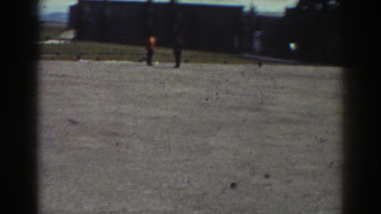 Two distant figures cross an open courtyard in front of a dark building.