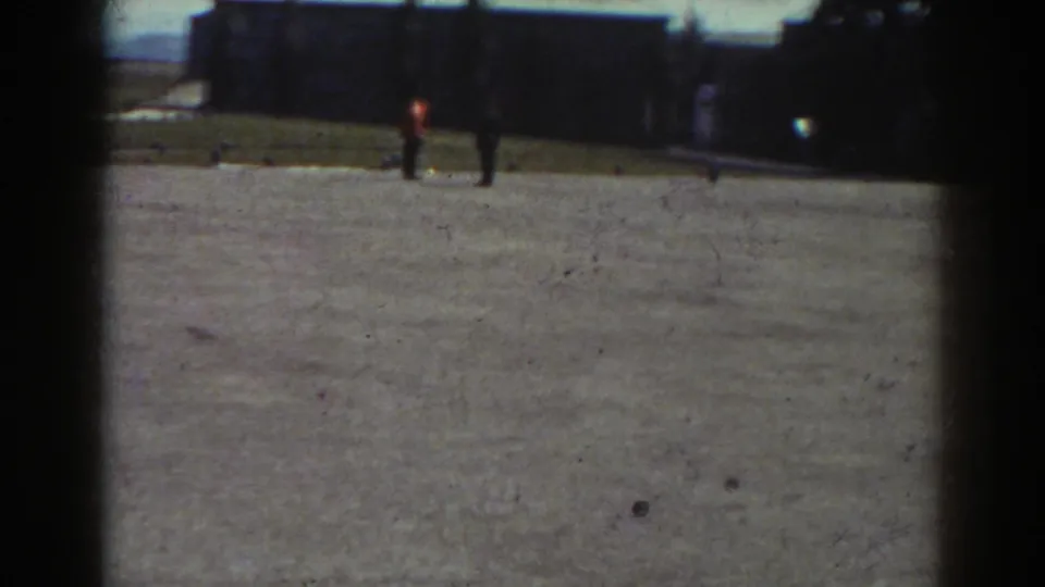 1938 Quebec frame with two distant figures walking across a pale gravel courtyard near dark historic-looking buildings.