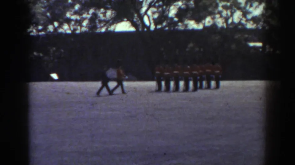 1938 Quebec frame showing two figures crossing near red-jacketed guards lined up on a courtyard under trees.
