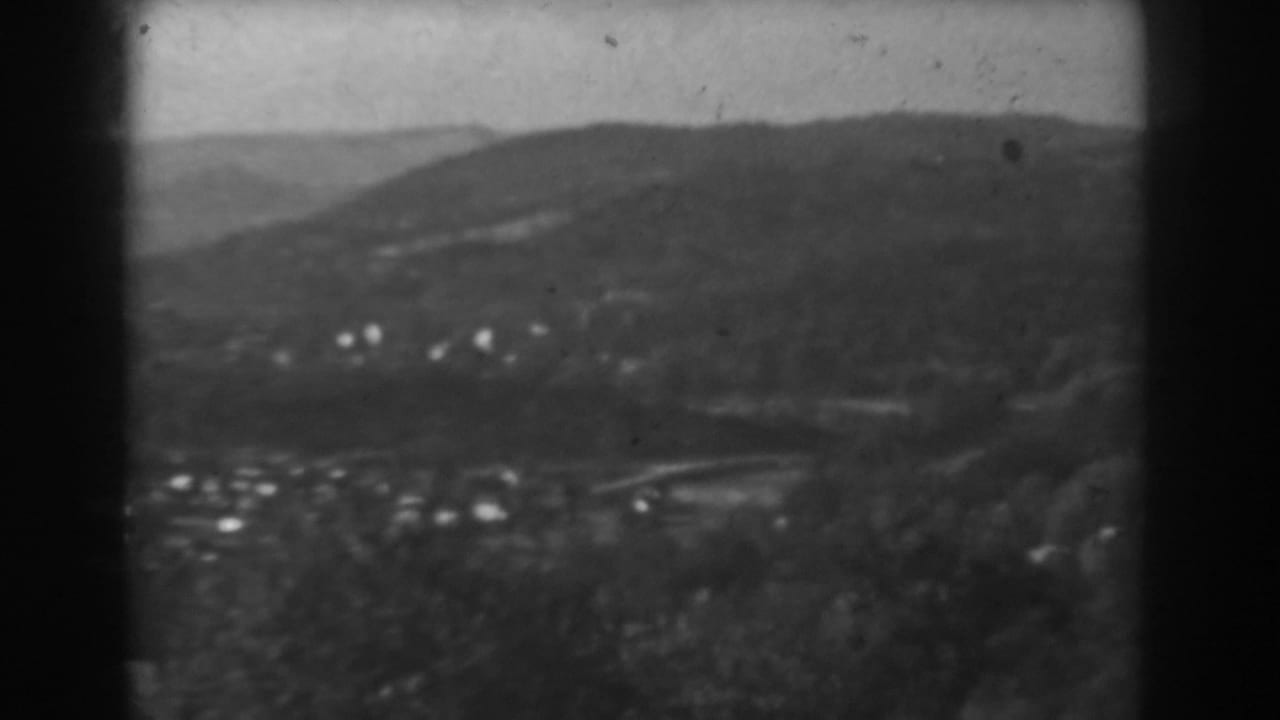Soft black-and-white view across a valley with wooded hills and scattered small houses.