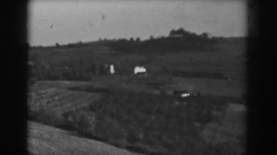 1938 Vineland frame showing two small white structures in a rural field with low hills behind them.