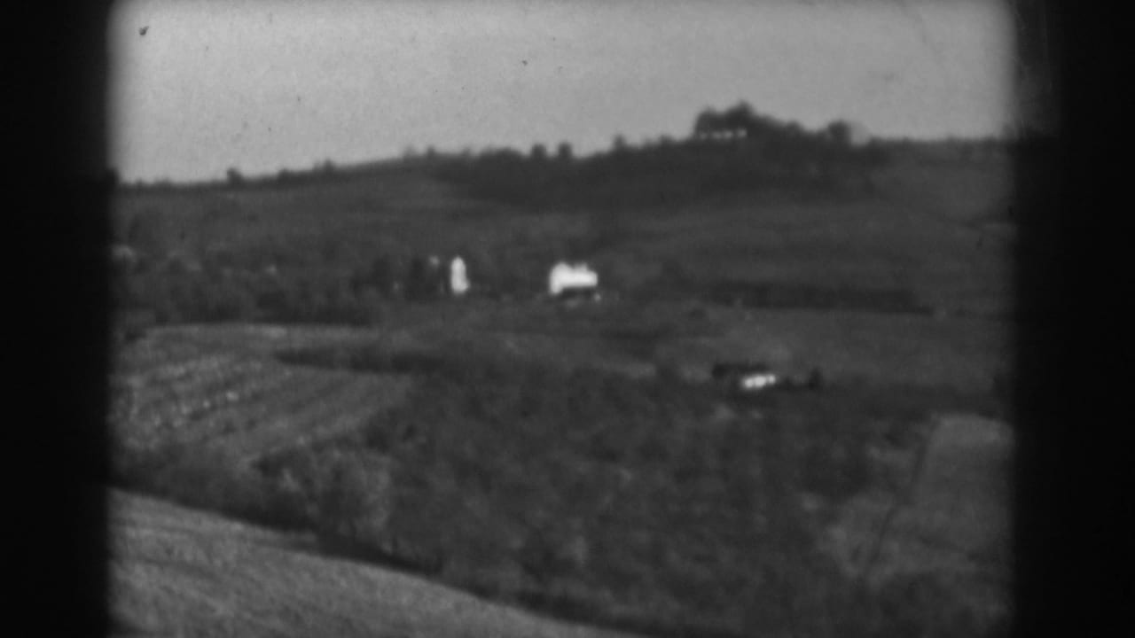 Soft black-and-white view of rolling fields, tree lines, and distant farm buildings.