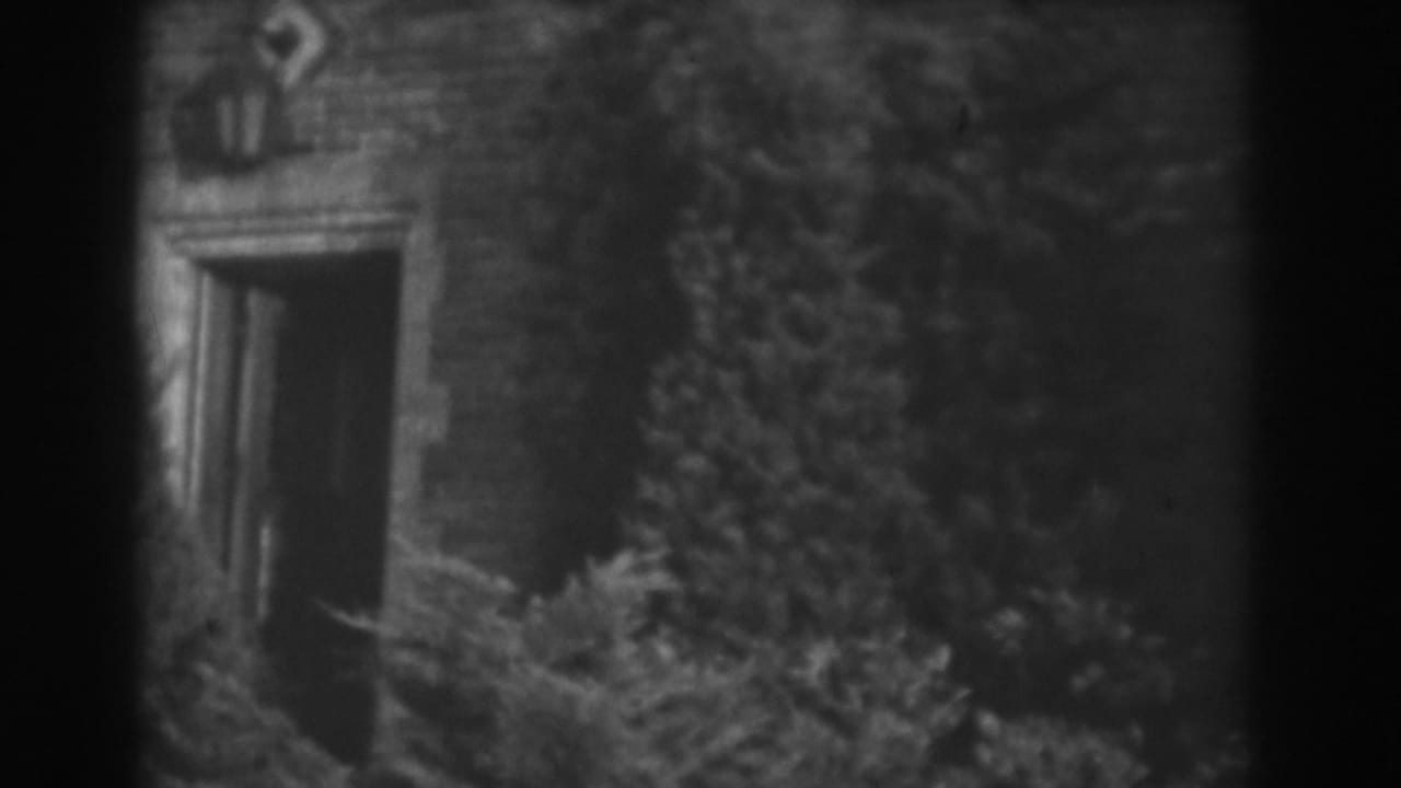 1938 Vineland home-movie frame showing a brick exterior, dark doorway, and overgrown vegetation along the wall.