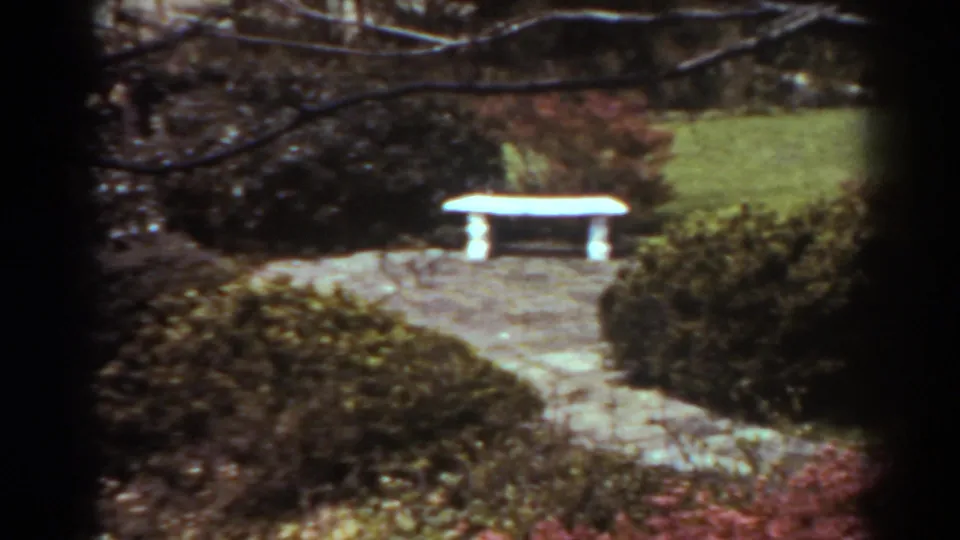 Vintage 8mm garden frame from West Virginia showing a white bench, stone walkway, lawn, and dense shrubs in reddish foliage.