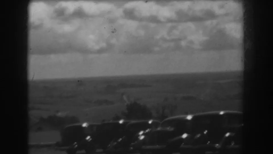 West Virginia home-movie frame with a row of 1930s automobiles below an open, clouded rural horizon.