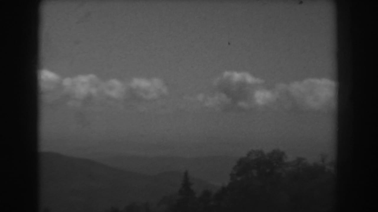Distant hills and tree silhouettes beneath a band of clouds in a black-and-white 8mm frame.