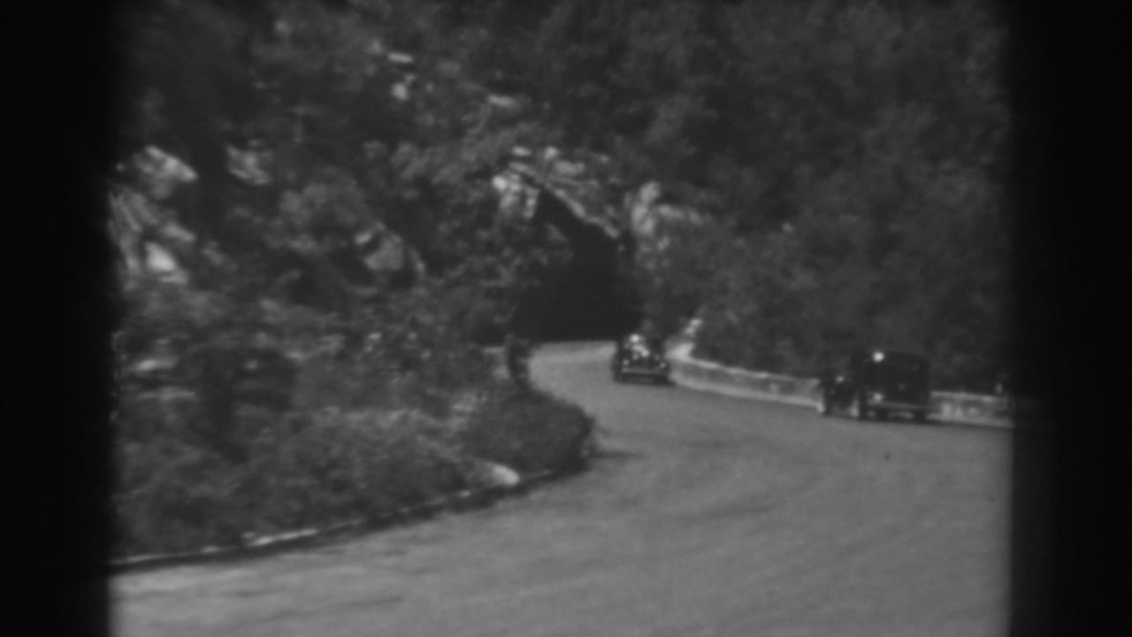 Two period cars appear on opposite sides of a winding West Virginia mountain road bordered by trees and a tunnel opening.
