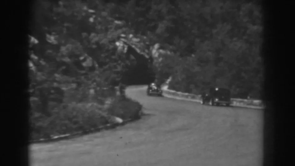 Black-and-white 8mm frame of cars on a curving West Virginia mountain road, with a dark tunnel mouth in the trees.