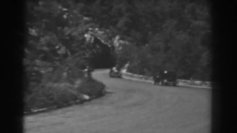 A roadside 8mm view shows two automobiles on a wooded mountain curve in West Virginia, with a tunnel entrance ahead.
