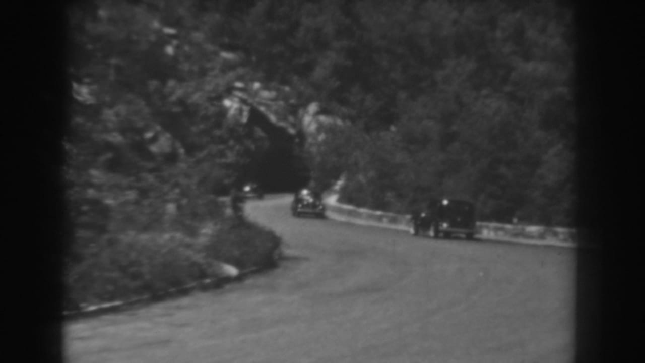 1938 West Virginia home-movie frame of cars on a winding mountain road approaching a dark tunnel beneath a wooded hillside.