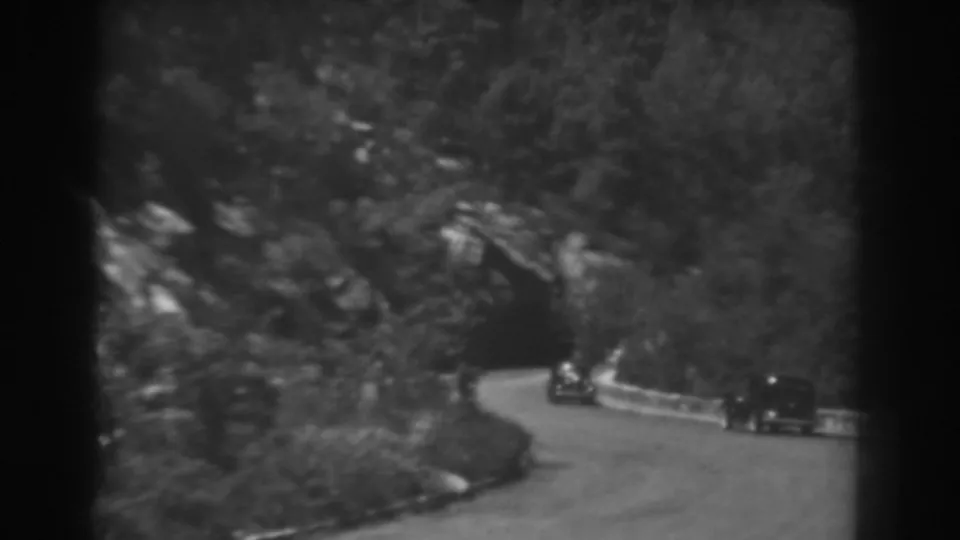 A home-movie frame shows one vintage car near the tunnel mouth and another following on the wooded mountain curve in West Virginia.