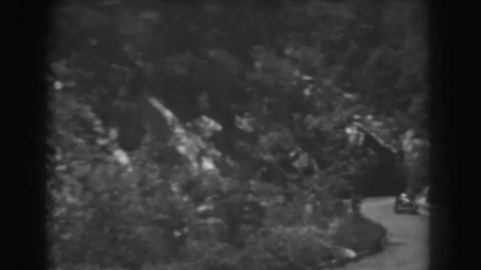 Late clip frame with a car near the edge of the view, a dark tunnel opening, dense trees, and rocky mountain terrain in West Virginia.
