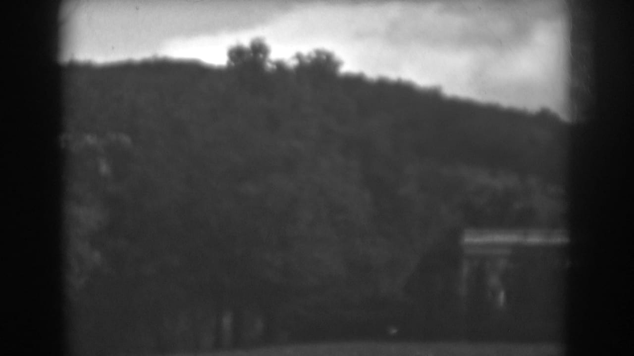 Black-and-white 8mm frame of a wooded hillside in Winchester, Maryland, with a small structure partly visible near the lower right edge.